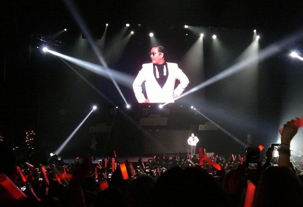 Psy in a white jacket on a large concert screen performing Gangnam Style, with the crowd in the foreground