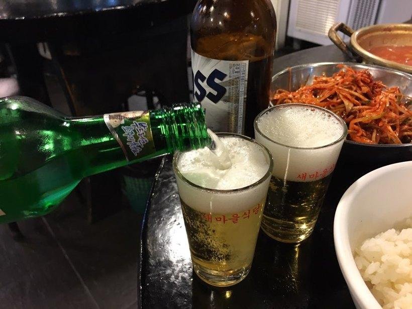 Soju being poured from a green bottle into a glass of beer, with a second glass and side dishes on the table