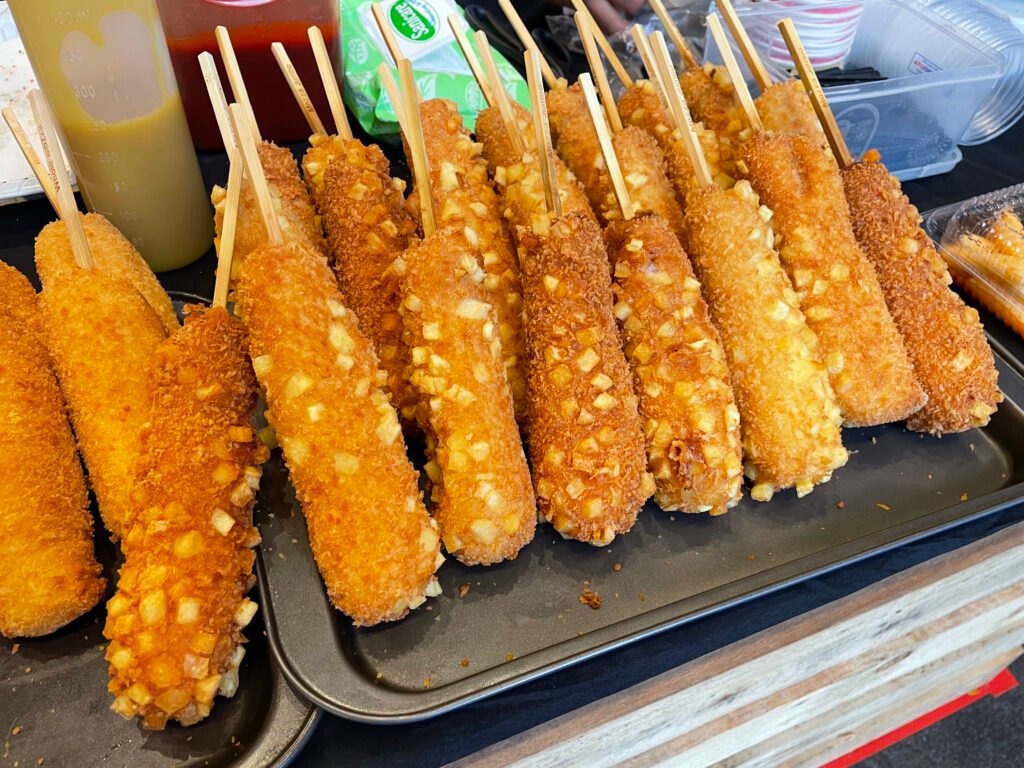 A Korean corn dog coated in batter and crumbs, held on a wooden stick